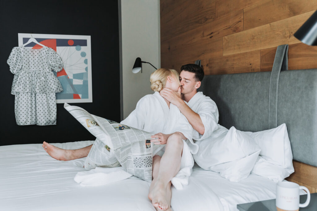 Bride and groom kiss on bed while holding newspaper and in their robes at their hotel