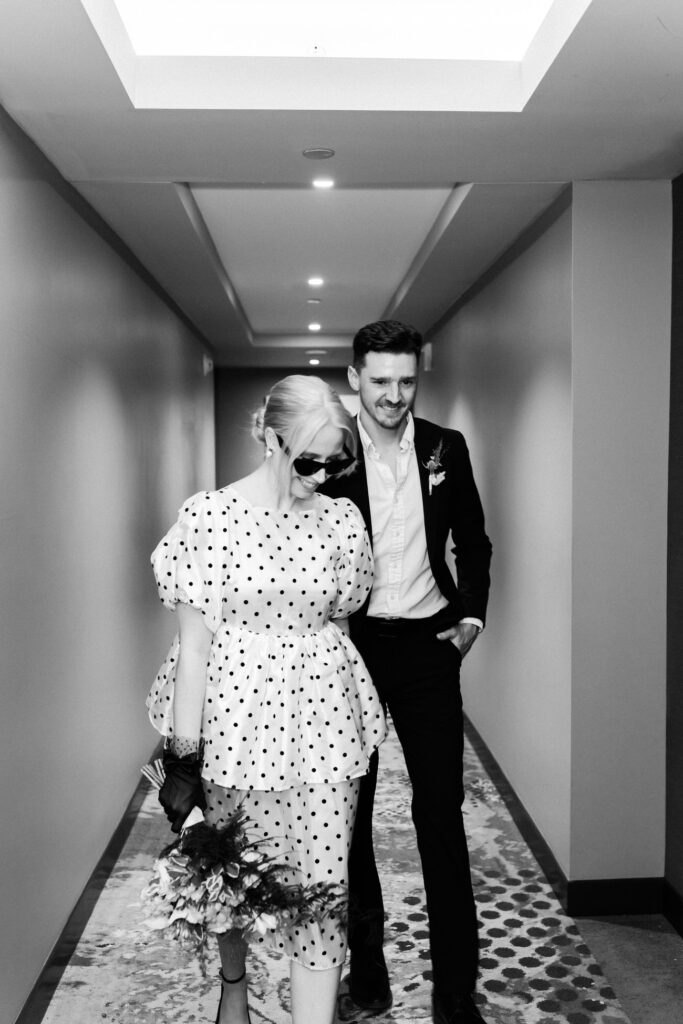Girl walks towards camera while holding guys hand. She is in a polka dot dress and wearing sunglasses