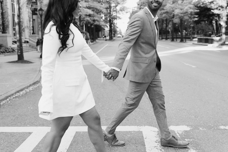 couple holds hands and walks through crosswalk of city.